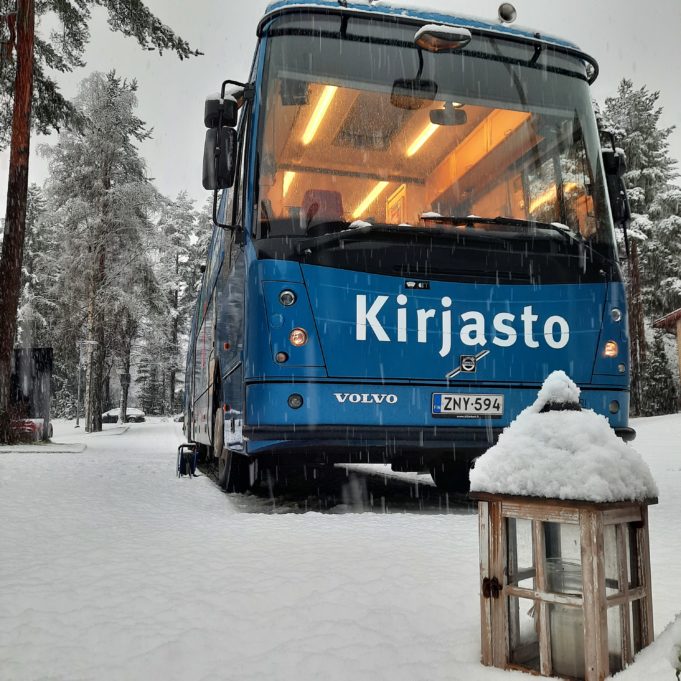 Kirjastoauto Kotikujan Konsta lumisessa maisemassa. Etualalla puinen lyhty.