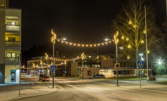 Katunäkymä pimeän aikaan, kadun varrella näkyy valopylväisiin kiinnitettyjä kausivaloja, pysäköityjä autoja ja rakennuksia.