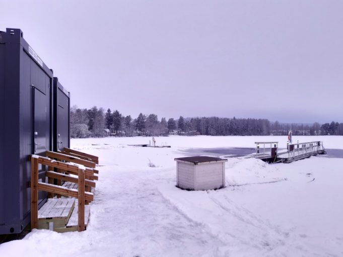 Talvinen rantamaisema, jossa näkyy myös laituri, metsän reunaa kauempana ja etualalla rannassa pukukopit.