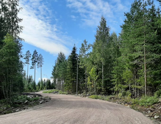 Ensi vasemmalle ja sitten oikealle kaartuva hiekkatie, jonka molemmilla puolilla näkyy kesäistä metsää. edessä näkyy sinivalkoinen taivas.