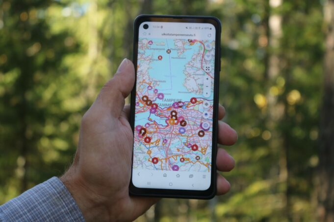 Käsi, jossa on matkapuhelin, ja puhelimen näytöllä näkyy kartta. Taustalla on metsää.