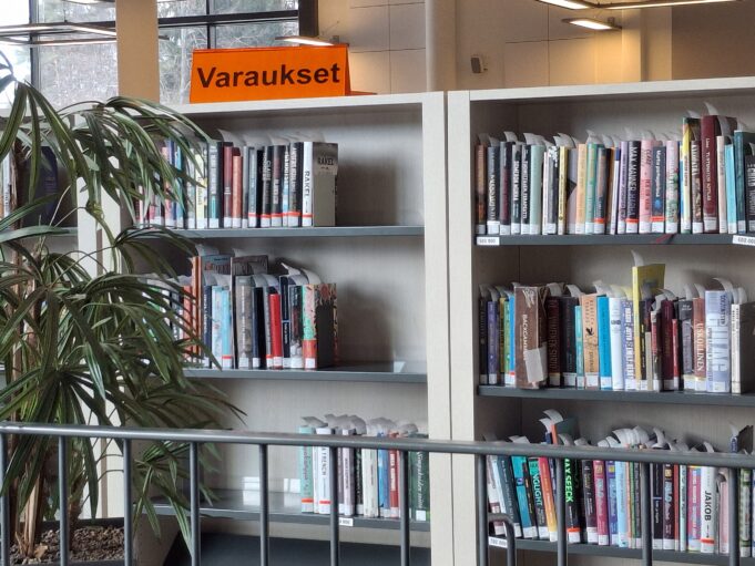 Kirjahyllyjä täynnä kirjoja, hyllyn päällä kyltti varaukset. Lisäksi viherkasvin oksia.