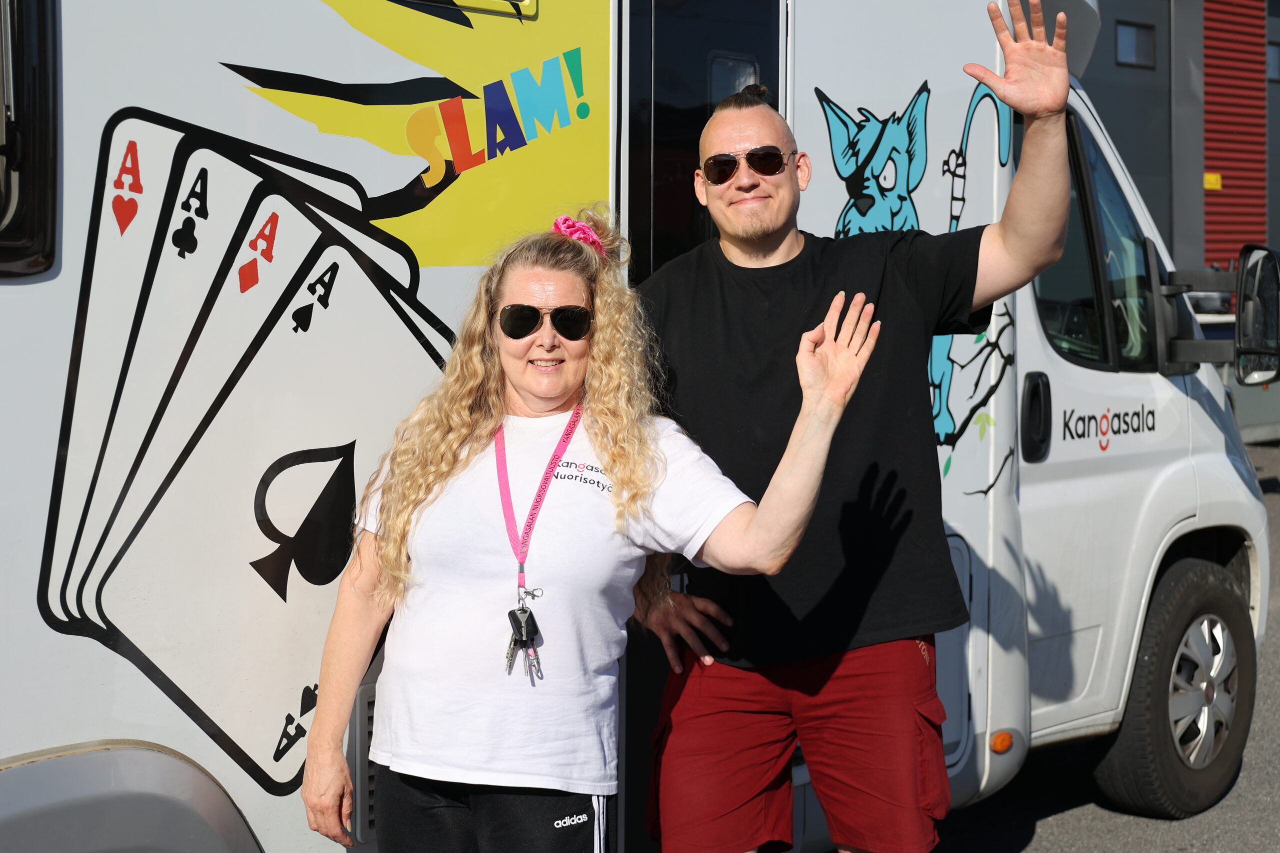 Kaksi aikuista seisoo liikkuvan nuorisotila-auto Kyläkolli, eli valkoisen matkailuauton edessä ja vilkuttaa aurinkolasit päässä.