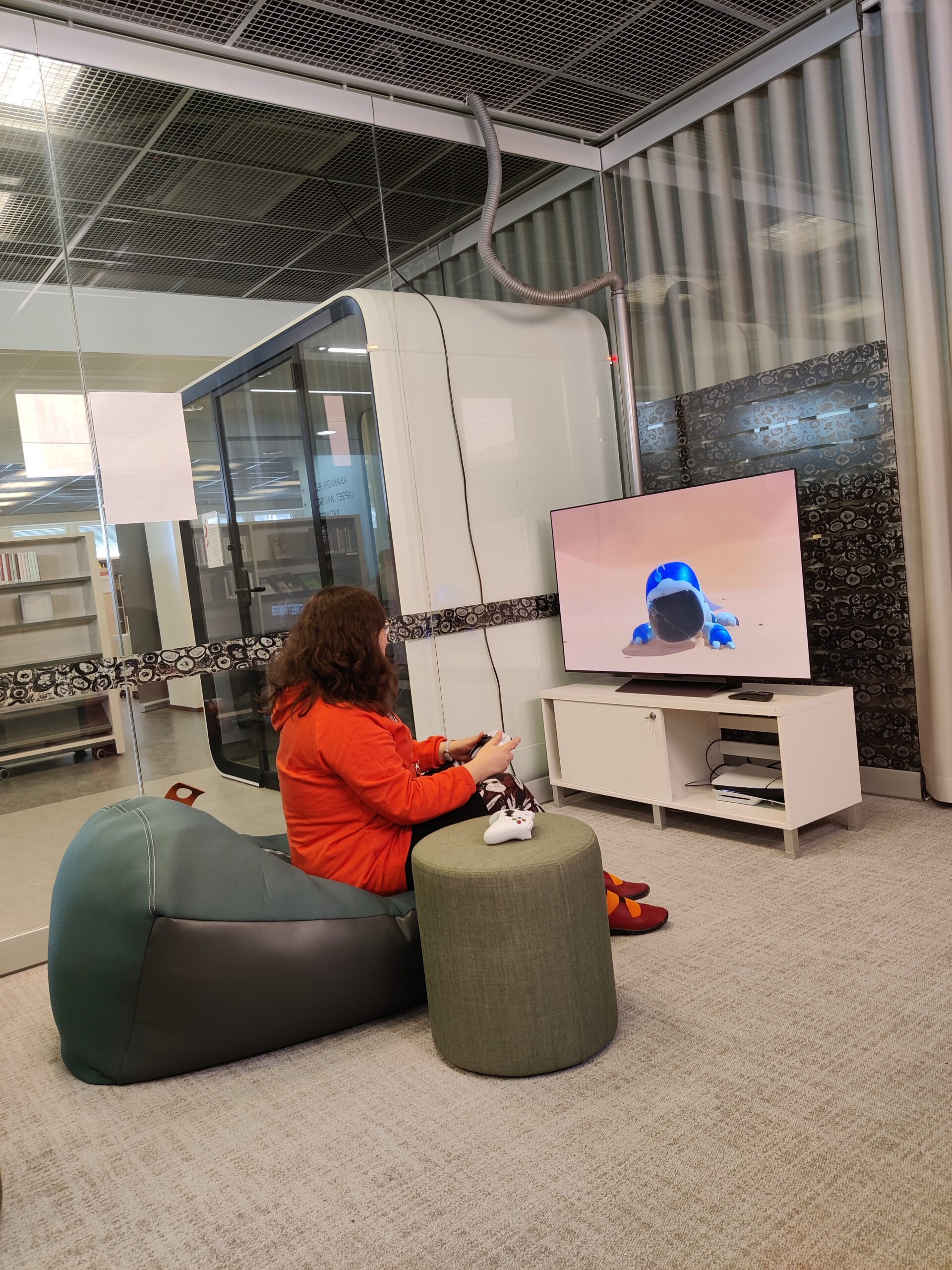 Pelitila, jossa punapaitainen nainen istuu vihreällä säkkituolilla ja pelaa konsolipeliä.