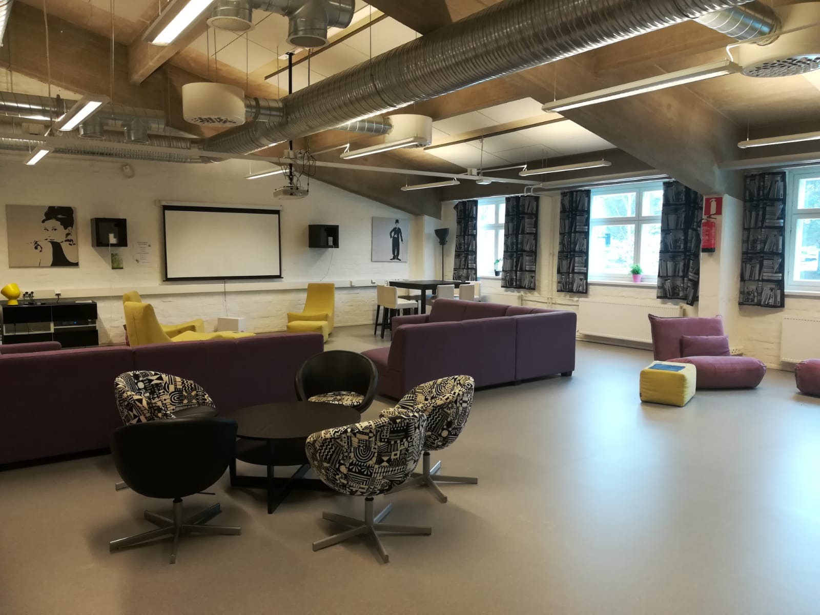 Kuvassa valkotiiliseinäinen huone, katossa ilmanvaihtoputkia. Etualalla matala pöytä ja neljä nojatuolia. Taustalla kaksi suurta violettia moduulisohvaa.