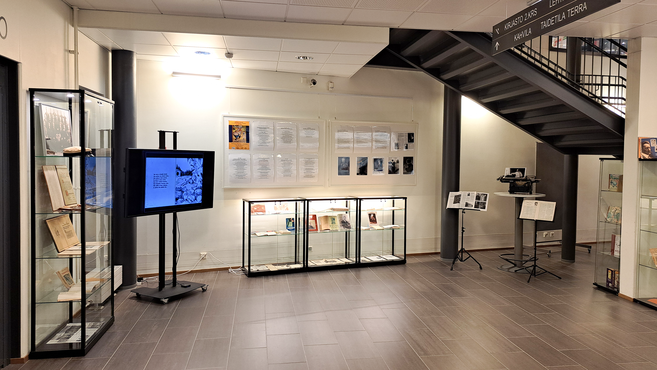 Portaikon alla lasiseinäisiä vitriinejä ja iso näyttö. Seinällä ripustuskisko.