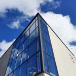 Alhaalta päin kuvattuna pääkirjaston lasiseinäinen kulma. Taustalla sininen taivas ja poutapilviä.