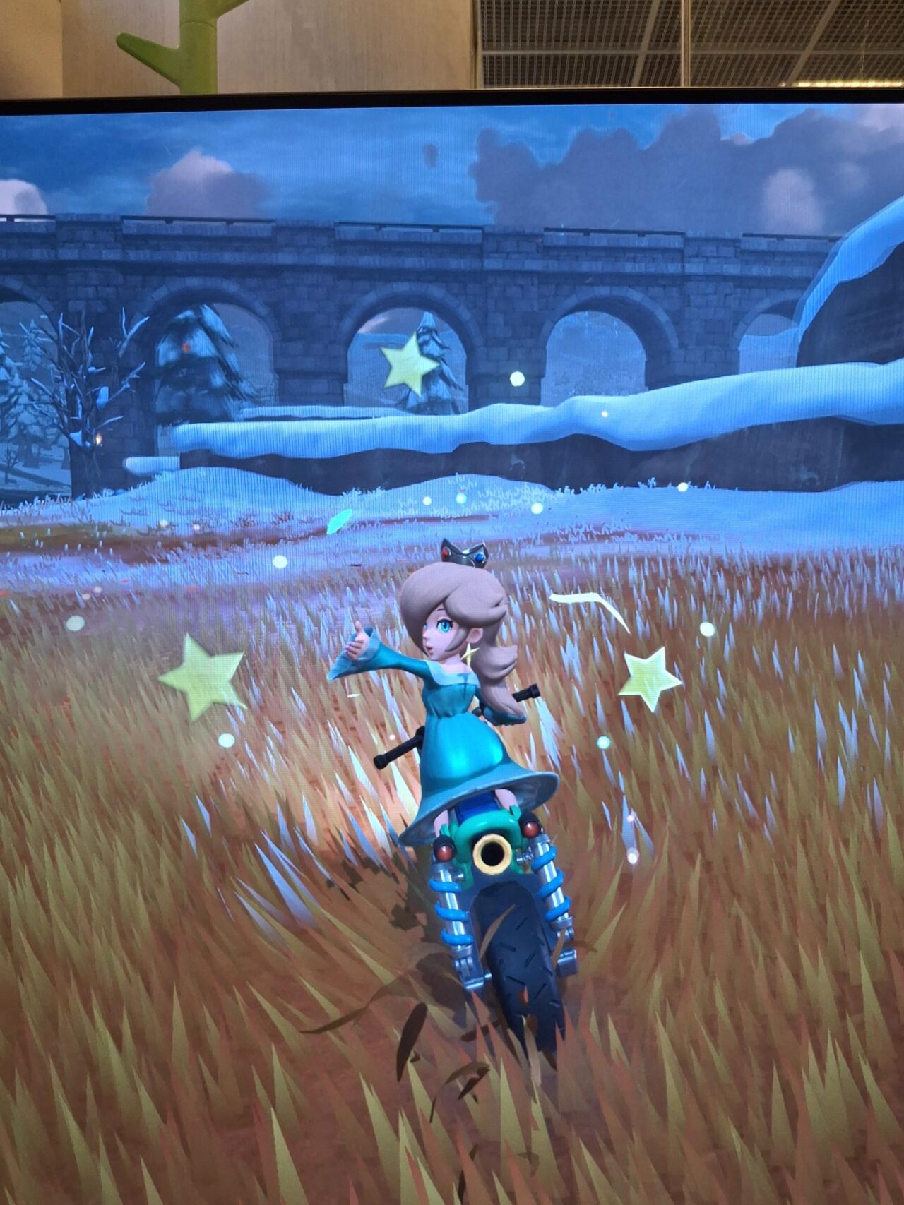 Konsolipelissä vihreäpukuinen naishahmo ajaa mopolla pellolla.