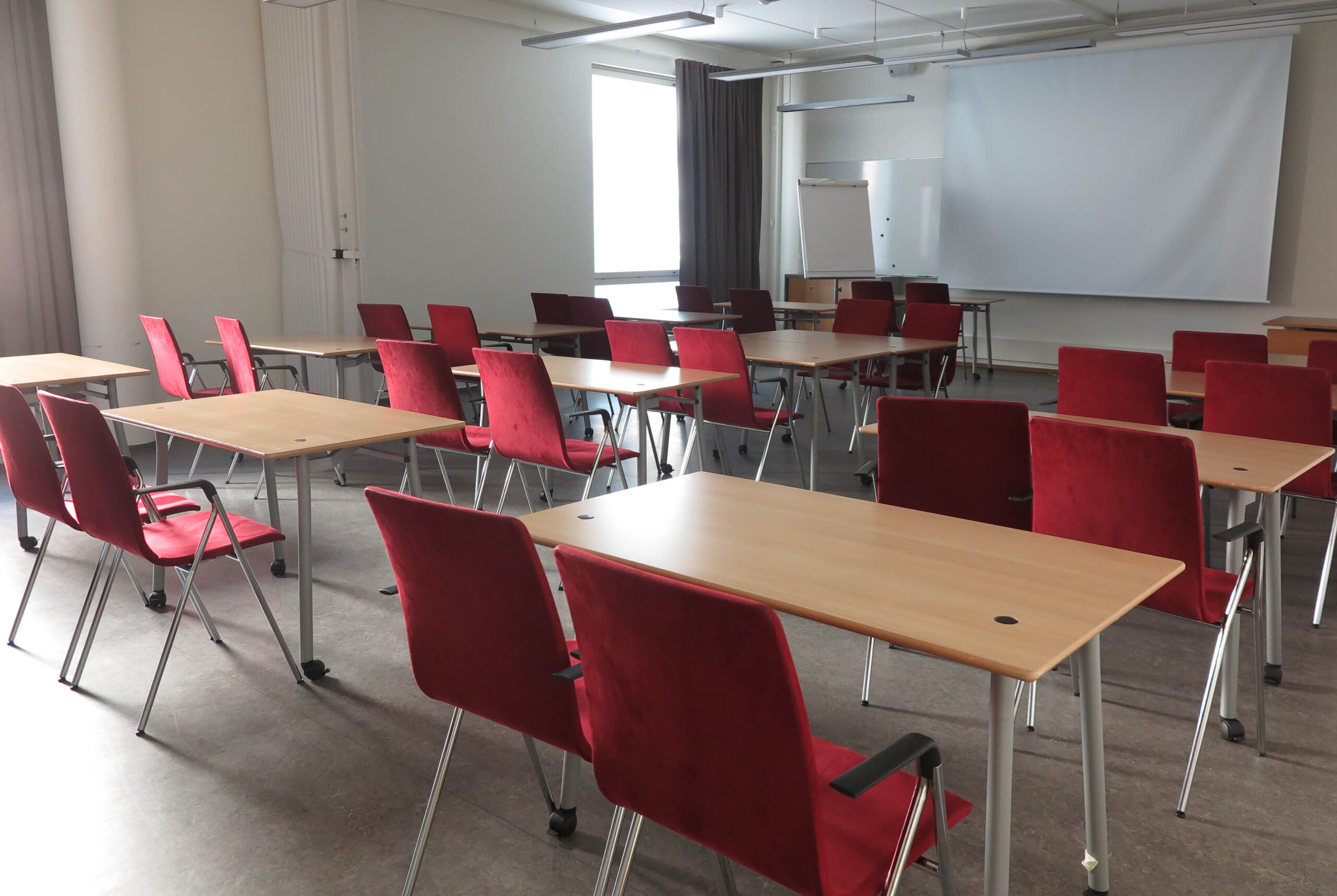 Iso sali, jossa pöytiä ja punaisia tuoleja ja salin edessä valkokangas esittämistä varten.