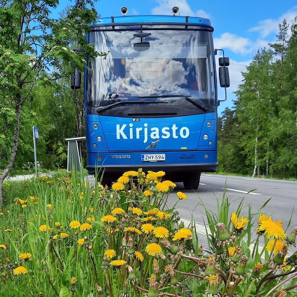 Kangasalan kirjastoauto Kotikujan Konsta kesäisessä maisemassa. Etualalla on keltaisia kukkia.
