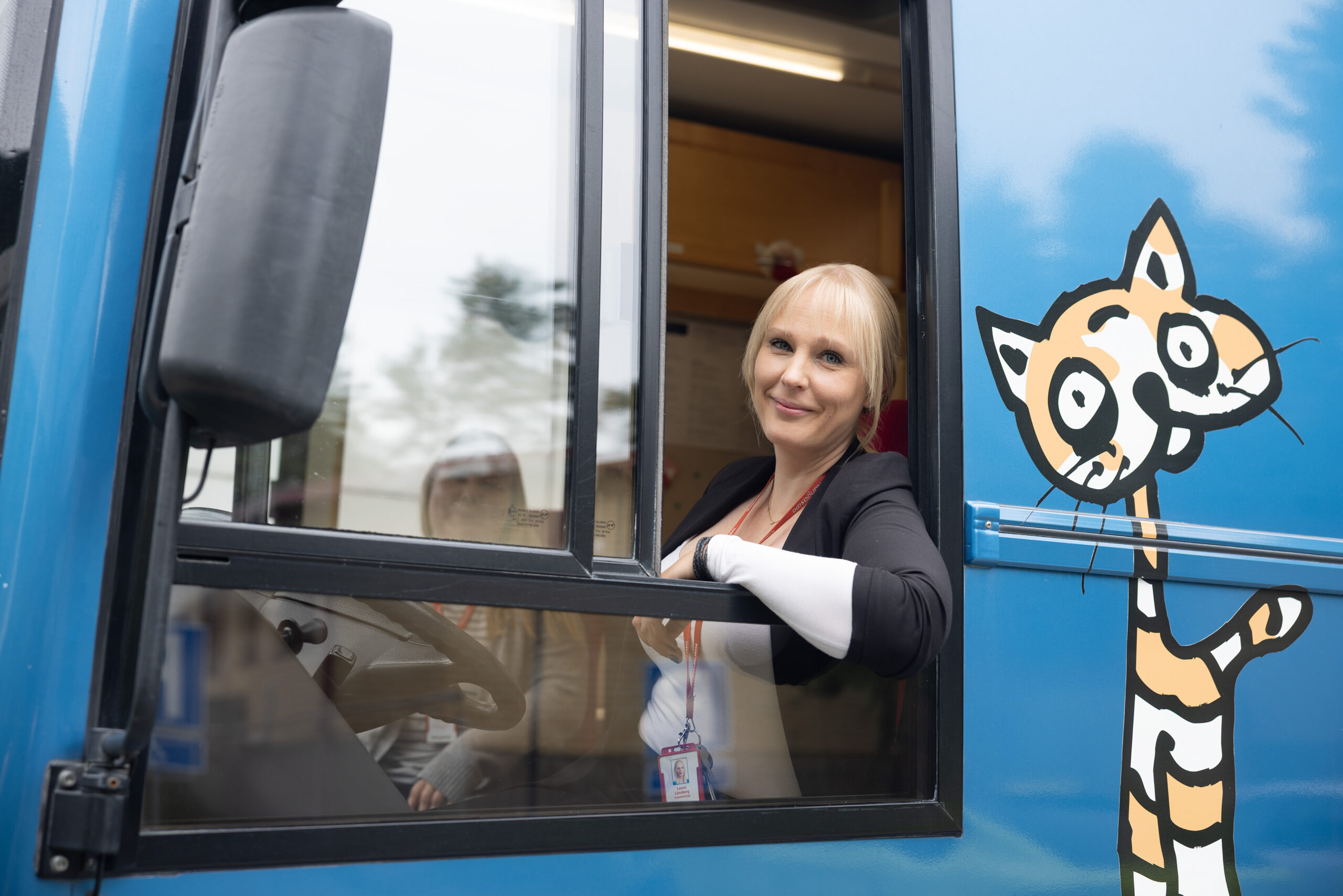 Kirjastoauton kuljettaja kurkkaa hymyillen avoimena olevasta kirjastoauto Kotikuja Konstan etuikkunasta.