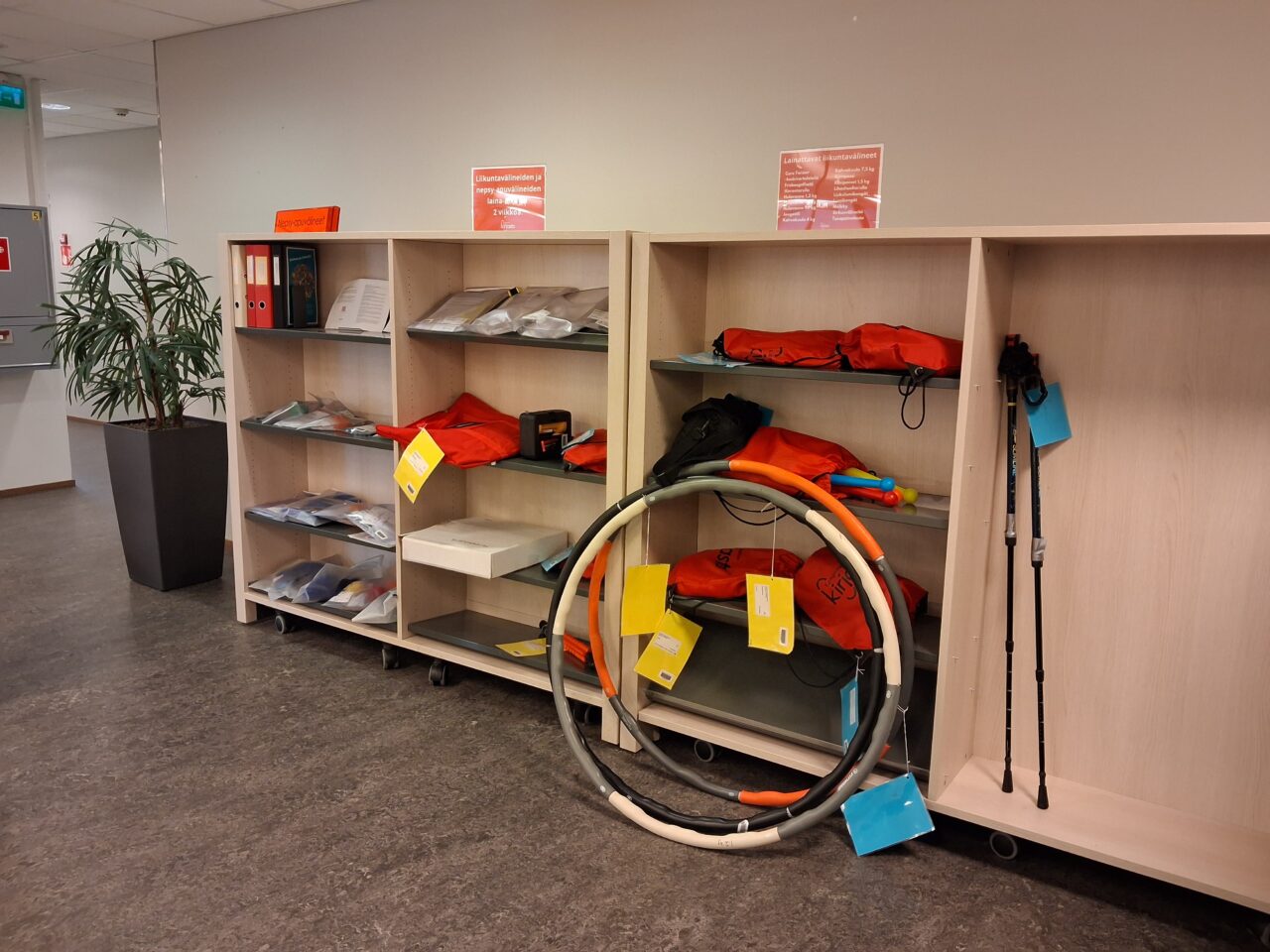 Vaaleita hyllyjä, joissa kirjastosta lainattavia liikuntavälineitä ja muita esineitä. Hyllyjen edessä niihin nojaamassa lainattavia hulavanteita.