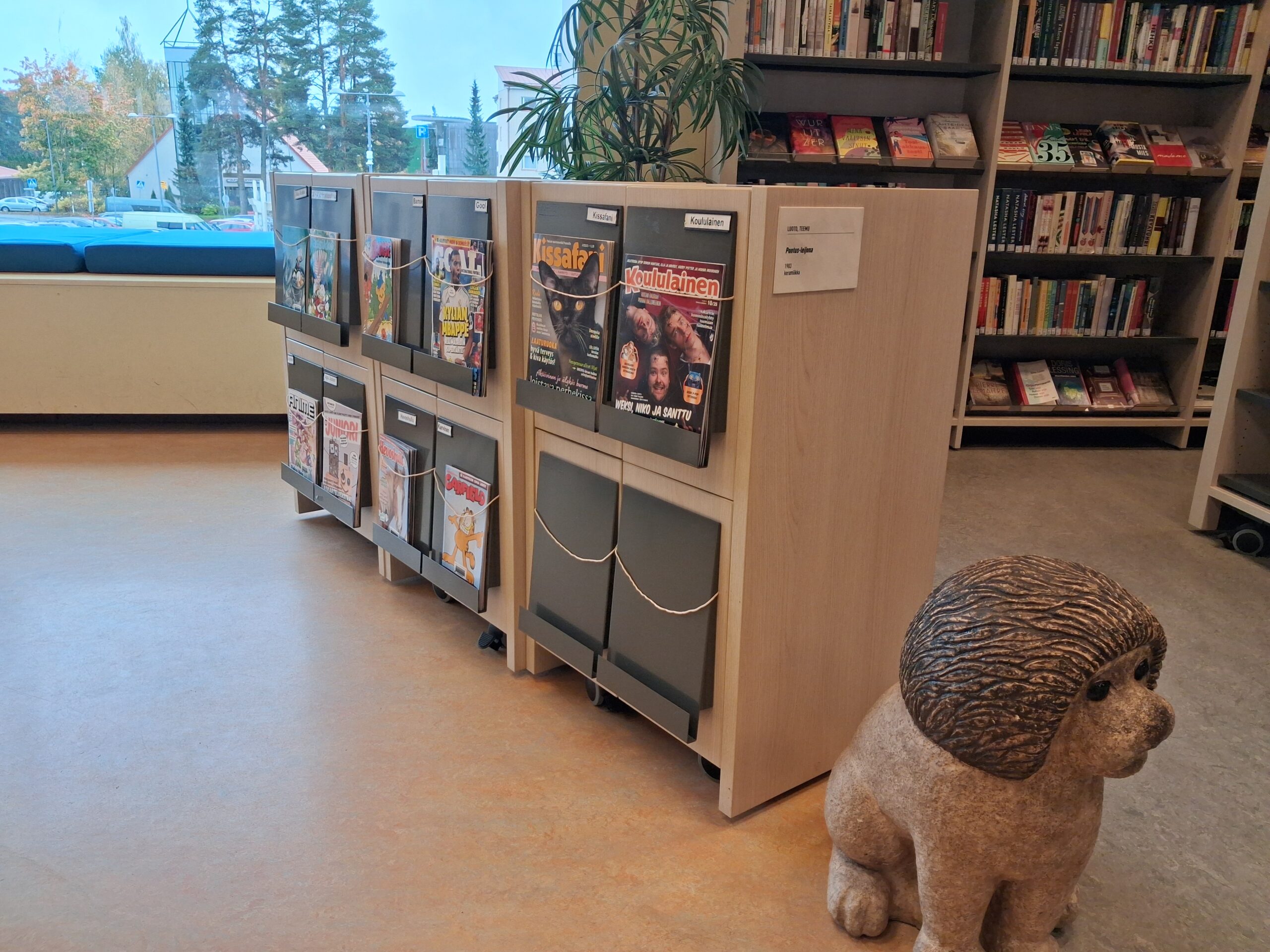 Matal hylly, jossa lapsille ja nuorille suunnattuja aikakauslehtiä. Vieressä leijonapatsas.