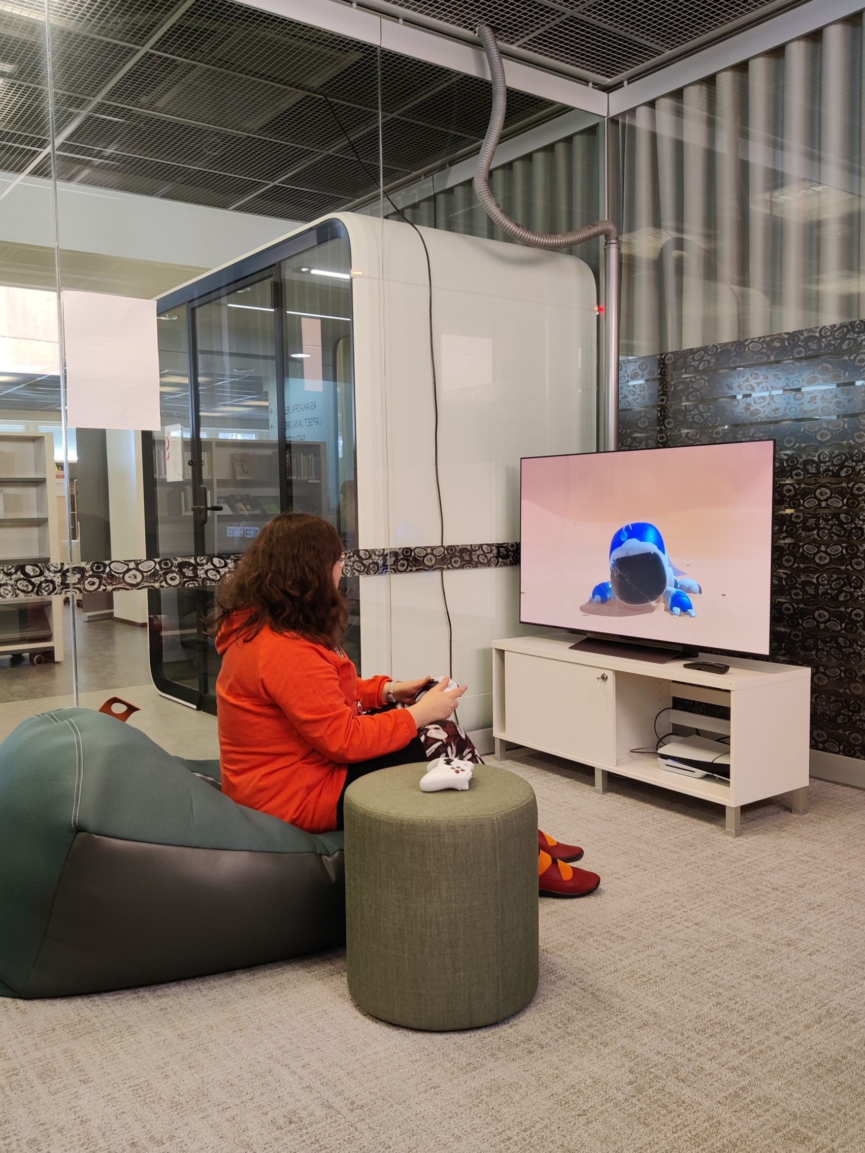 Pelitila, jossa punapaitainen nainen istuu vihreällä säkkituolilla ja pelaa konsolipeliä.