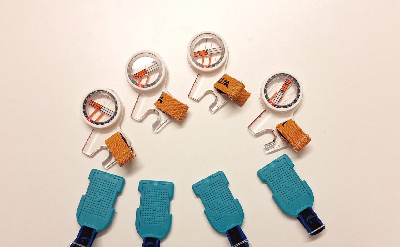 Valkoisella pöydällä neljä suunnistuksen peukalokompassia ja emit-korttia ajanottoon.