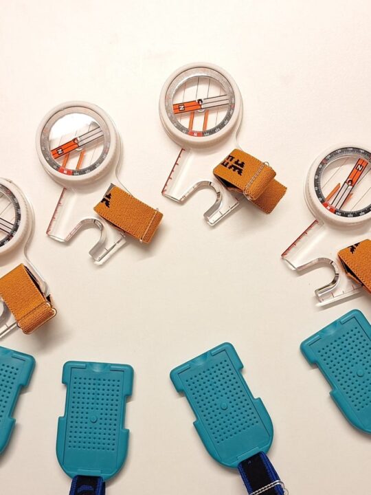 Valkoisella pöydällä neljä suunnistuksen peukalokompassia ja emit-korttia ajanottoon.