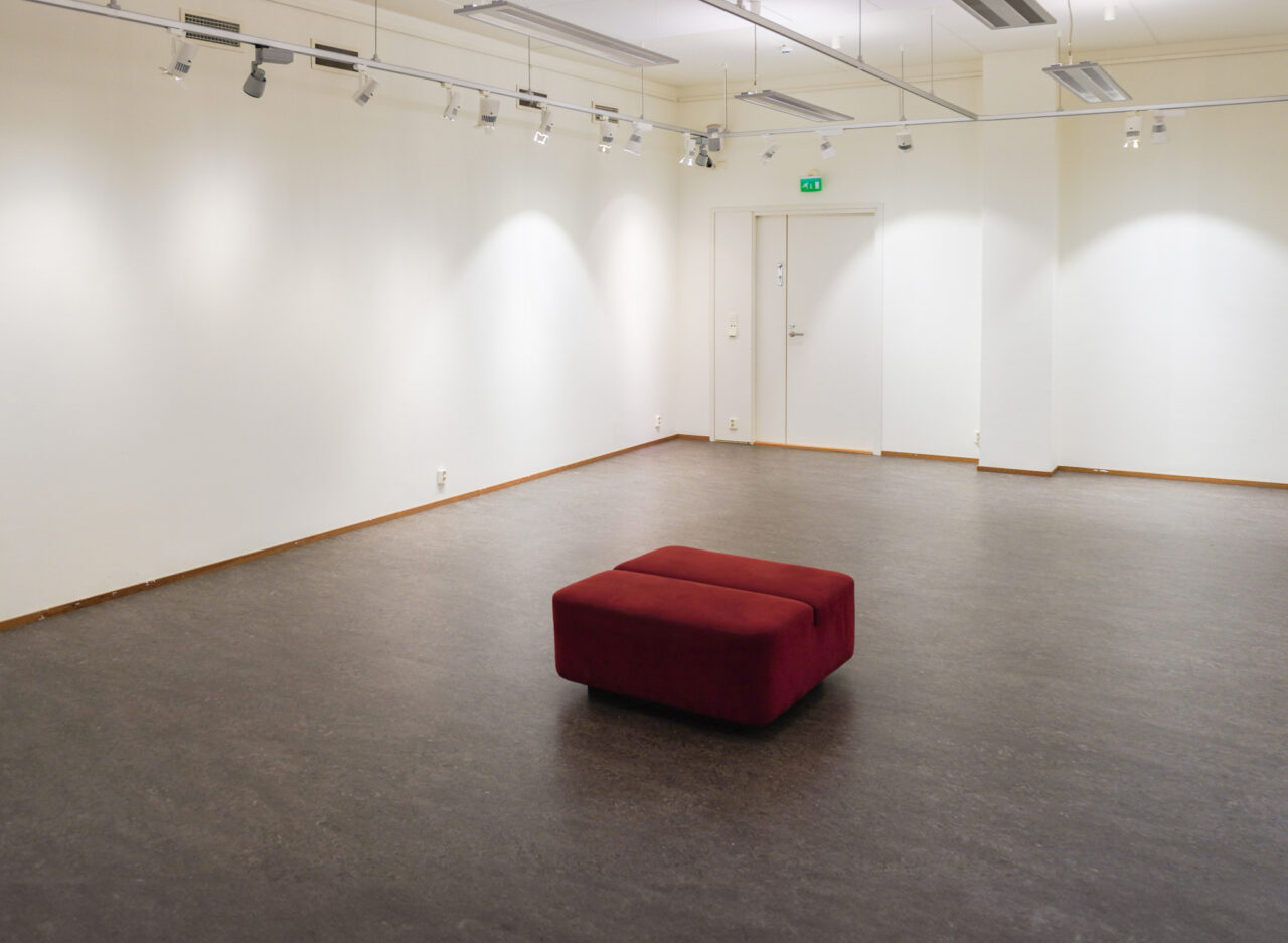 Tyhjä valkoseinäinen näyttelytila Terra. Keskellä huonetta viininpunainen rahi.