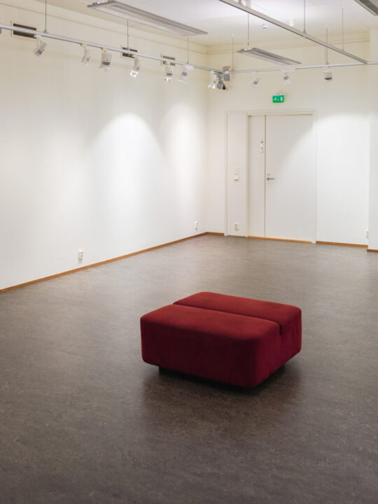 Tyhjä valkoseinäinen näyttelytila Terra. Keskellä huonetta viininpunainen rahi.