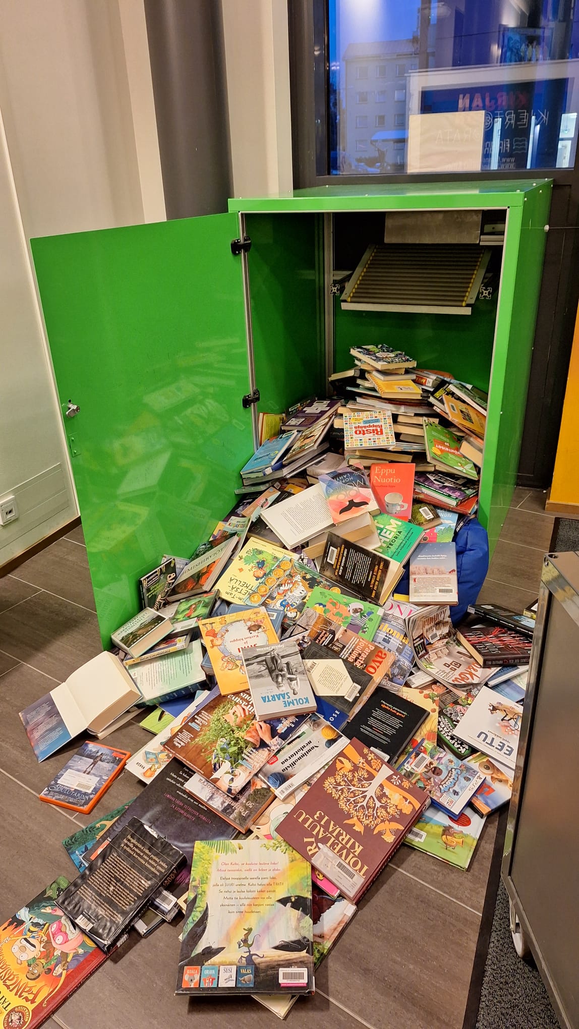 Vihreä palautuslaatikko avattuna. Sieltä vyöryy palautettuja kirjoja pitkin lattiaa. 