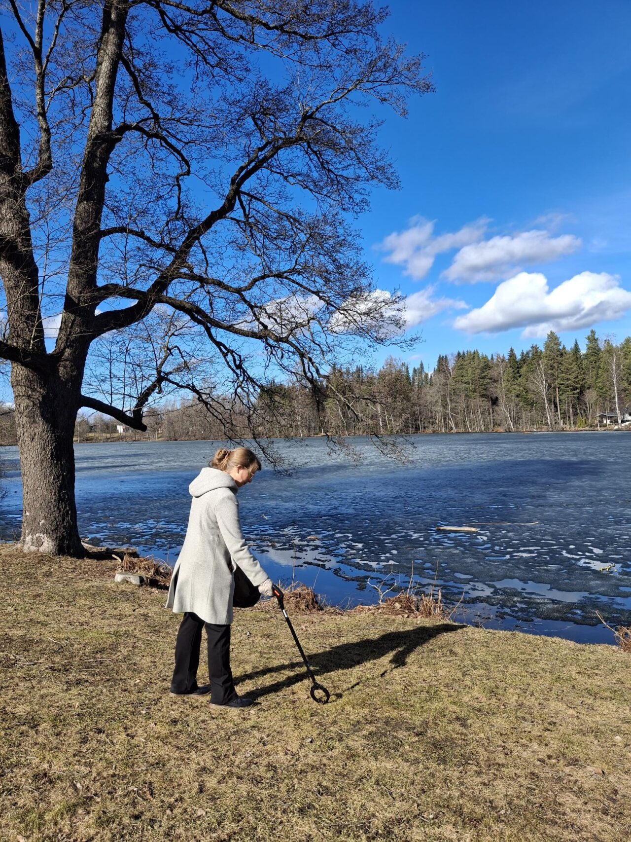 Nainen poimii roskia roskapihdeillä aurinkoisena kevätpäivänä järven rannalla.
