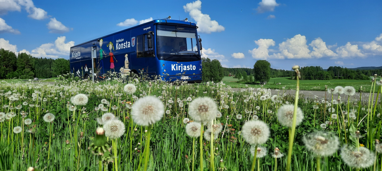 Kangasalan kirjastoauto kesäisessä maisemassa pellon reunassa. Pellolla kasvaa voikukkaa. Etualalla on pörröisiä voikukkia valmiina levittämään siemenet.