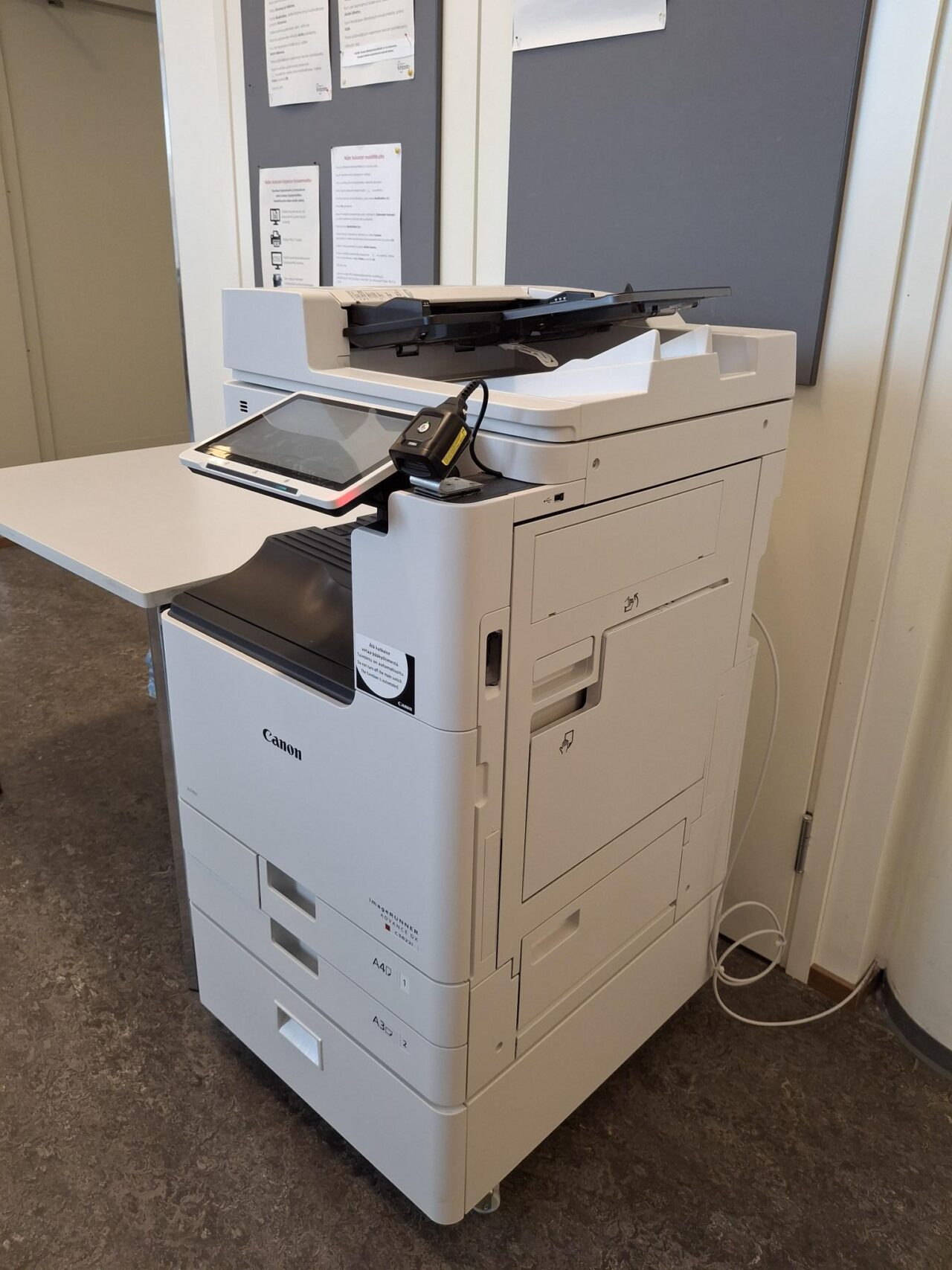 Kopiointiin, tulostamiseen ja skannaukseen tarkoitettu monitoimilaite seinän vieressä.