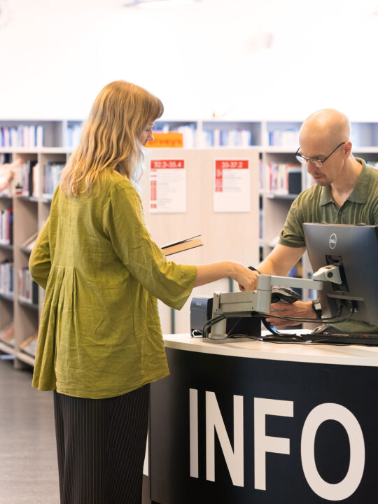 Kirjaston infotiskin äärellä kirjaston työntekijä ja asiakas. Taustalla kirjaston kirjahyllyjä.