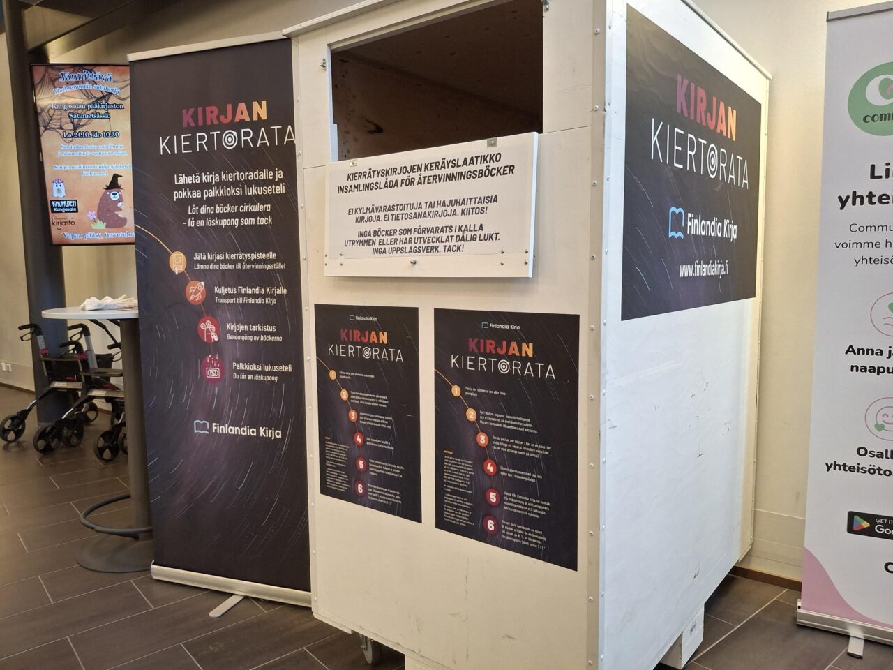 Kirjaston aulassa iso, valkoinen kirjankierrätyskontti sekä Finlandia Kirjan mainoksia.