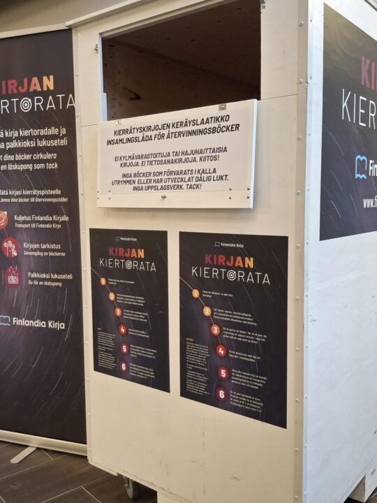 Kirjaston aulassa iso, valkoinen kirjankierrätyskontti sekä Finlandia Kirjan mainoksia.